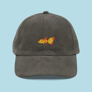 May include: A dark grey corduroy baseball cap with an embroidered cartoon goldfish in yellow, orange, and red. The cap has a curved peak and a small button on top. The background is light blue.