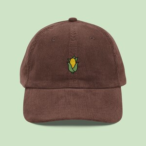 Op de afbeelding: Bruine corduroy baseballcap met een geborduurd maïsontwerp in geel en groen. De cap heeft een gebogen klep en een verstelbare band. Een casual en modieus accessoire.