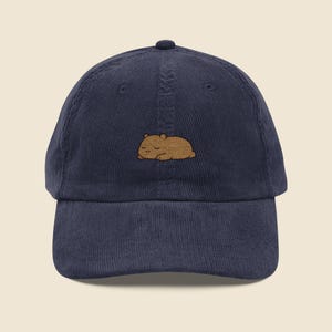May include: Navy blue corduroy baseball cap with a small embroidered brown capybara. The cap has a curved peak and an adjustable strap. A casual accessory for everyday wear, perfect for animal lovers.