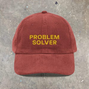 May include: A maroon corduroy baseball cap with a curved brim. The cap features the embroidered text "PROBLEM SOLVER" in a yellow color across the front. The cap has a rounded top and an adjustable strap.