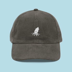 May include: An olive green corduroy baseball cap featuring a white embroidered squid design. The cap has a curved peak and an adjustable strap. A fashionable accessory for everyday wear.