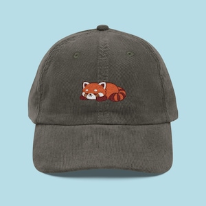 May include: A green corduroy baseball cap with an embroidered red panda design. The red panda is depicted lying down, with a white face, red and brown fur, and a striped tail. The cap has a curved peak and a button on the crown.