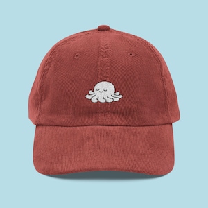 May include: A brick-red corduroy baseball cap featuring a white embroidered octopus design. The octopus has a smiling face. The cap has a curved peak and an adjustable strap at the back.