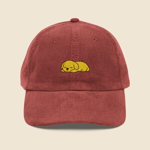 May include: A deep red corduroy baseball cap with a small, embroidered image of a sleeping yellow dog. The cap has a curved peak and an adjustable strap for a comfortable fit.