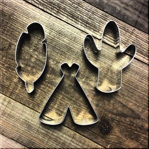 May include: Three metal cookie cutters in the shape of a cactus, a teepee, and a feather. The cutters are on a wooden surface.