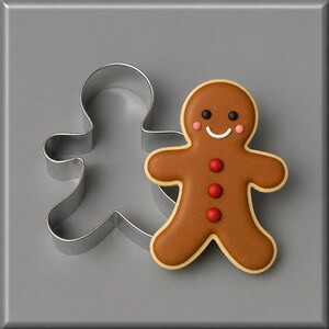 May include: A silver metal gingerbread man cookie cutter next to a decorated gingerbread man cookie. The cookie has brown icing, a white smile, black eyes, pink cheeks, and three red circular buttons down the center of its torso.