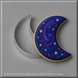 May include: A crescent moon-shaped metal cookie cutter next to a crescent moon-shaped cookie decorated with a blue and purple galaxy swirl pattern and small silver star sprinkles. The text AmericanTraditionCookieCutters.com is at the bottom.
