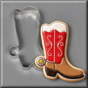 May include: A silver metal cowboy boot cookie cutter next to a decorated sugar cookie shaped like a cowboy boot. The cookie features a red shaft with white scrollwork, a tan foot, a dark brown heel, and a yellow star-shaped spur detail.