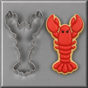 May include: A silver metal lobster-shaped cookie cutter next to a matching lobster-shaped cookie decorated with smooth red icing, raised red details, and two small black dots for eyes on a light-colored, plain, neutral background.