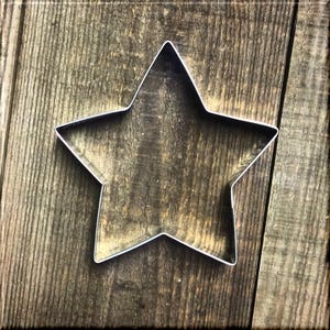 May include: A silver-colored star-shaped cookie cutter. The cutter is made of metal and has a simple, clean design. The background is a rustic wooden surface with visible grain.