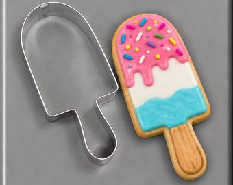 Puede incluir: Un cortador de galletas de metal con forma de helado junto a una galleta decorada con forma de helado. La galleta presenta un mango marrón, una sección central azul y una capa superior rosa cubierta con chispas de colores.