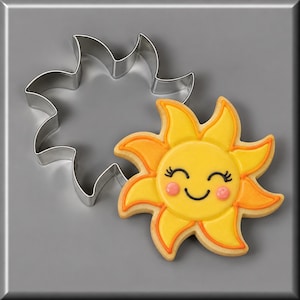 May include: A metal sun-shaped cookie cutter next to a matching sun-shaped cookie decorated with yellow and orange icing, featuring a smiling face with closed eyes, black eyelashes, and pink cheeks, all rendered in a bright, cheerful design.