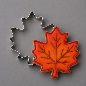 May include: A silver metal maple leaf cookie cutter positioned next to a maple leaf shaped cookie decorated with orange and red icing. The cookie features a raised red piped outline and central vein details, matching the shape of the metal cutter.