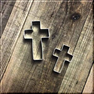 May include: Two metal cookie cutters in the shape of crosses. The larger cross is slightly wider than the smaller cross. Both crosses are silver in color and are sitting on a wooden surface.