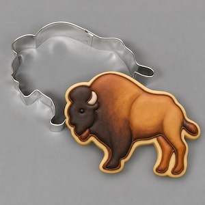 May include: A silver metal cookie cutter shaped like a bison, paired with a decorated sugar cookie in the same bison shape. The cookie features detailed icing in shades of dark brown, light brown, and cream, depicting the animal's profile.