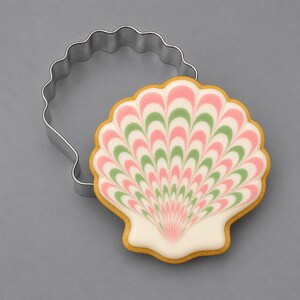 May include: A silver metal seashell-shaped cookie cutter positioned next to a matching seashell-shaped biscuit decorated with white icing and a feathered pattern of alternating pink and green lines radiating from the base to the scalloped edge.