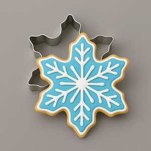 May include: A metal snowflake-shaped cookie cutter positioned behind a snowflake-shaped cookie decorated with light blue icing and white piped lines. The cookie features a central star pattern with branching arms extending to the edges.