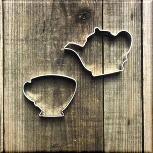 May include: Two metal cookie cutters, one shaped like a teapot and the other shaped like a teacup, on a wooden background.