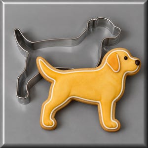 May include: A silver metal dog-shaped cookie cutter positioned next to a matching dog-shaped cookie decorated with yellow icing, white piped outlines, and a small dark brown dot for the eye and nose, creating a clear visual representation.