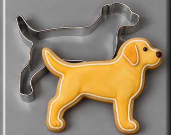 May include: A silver metal dog-shaped cookie cutter positioned next to a matching dog-shaped cookie decorated with yellow icing, white piped outlines, and a small dark brown dot for the eye and nose, creating a clear visual representation.