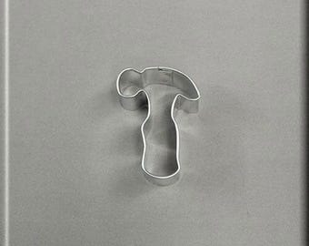 May include: A silver metal cookie cutter shaped like a hammer. The cookie cutter is on a grey background.