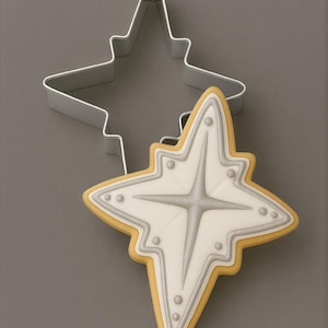 May include: A metal star-shaped cookie cutter and a matching star-shaped cookie decorated with white and silver icing. The cookie features a central four-pointed star design with small raised dots along the edges and a light tan border.