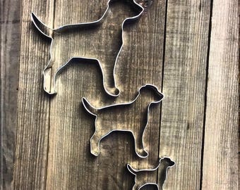 May include: Three metal dog cookie cutters of different sizes. The largest cutter is a silhouette of a standing dog, the middle cutter is a smaller silhouette of a standing dog, and the smallest cutter is a silhouette of a sitting dog.