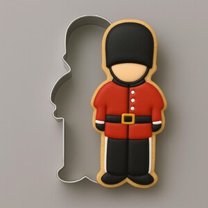 May include: A metal cookie cutter shaped like a British guard alongside a matching cookie decorated with red, black, and white icing. The cookie features a tall black bearskin hat, a red tunic with white buttons, a black belt with a gold buckle, and black trousers.
