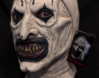 May include: A white clown mask with black eyes, red lips, and a black top hat. The mask has a distressed look and is mounted on a wooden base.