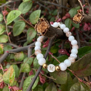 May include: White beaded bracelet with a gold letter "M" charm. The bracelet is on a branch with green leaves.
