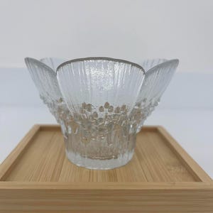 Peut inclure: Un bol en verre transparent avec un design texturé en forme de pétales. Le bol repose sur un plateau en bois brun clair. Le bord du bol a un bord lisse et la surface texturée crée un intérêt visuel.