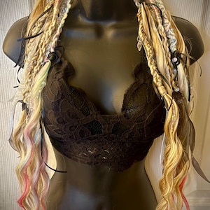 Può includere: Un bralette in pizzo nero è esposto su un manichino, abbinato a extension per capelli intrecciati biondi e castano chiaro. Le extension sono decorate con nastri neri, piume e ciocche di capelli colorati.