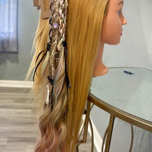 Könnte beinhalten: Ein Schaufensterpuppenkopf mit langem, blondem Haar, das mit einem seitlichen Zopf, einer Haarspange und farbenfrohen Akzenten gestylt ist. Der Zopf hat dekorative Charms und schwarze Bänder. Die Haarspitzen sind mit rosa Highlights versehen.