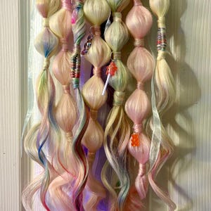 Könnte beinhalten: Ein skurriles Haaraccessoire mit mehreren Strähnen pastellfarbenen Haares, das mit bauchigen Abschnitten versehen und mit kleinen Charms verziert ist. Das Haar hat Strähnen in Rosa, Blau und Grün, mit Bändern und Accessoires.