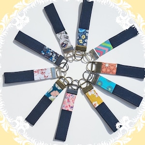 Peut inclure: Une collection de porte-clés avec des lanières en tissu bleu foncé et des accents en tissu à motifs. Les porte-clés présentent divers motifs, notamment floraux, géométriques et abstraits. Chaque porte-clés est doté d'un anneau métallique pour attacher les clés.