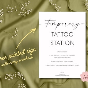 Pode incluir: Uma placa branca com a frase "Temporary Tattoo Station" em texto preto. As instru&ccedil;&otilde;es para aplicar tatuagens tempor&aacute;rias est&atilde;o listadas. Uma seta amarela aponta para o texto "Free printed sign with every purchase".