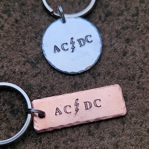 Może przedstawiać: Dwa breloki z logo zespołu AC/DC. Jeden brelok jest srebrny z okrągłą zawieszką, a drugi miedziany z prostokątną zawieszką.