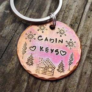 以下が含まれることがあります： 小屋のデザイン、雪片、ハートマークで「CABIN KEYS」と書かれた、銅色の丸いキーホルダーです。