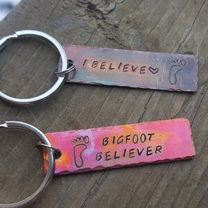 Op de afbeelding: Twee metalen sleutelhangers met een zilveren sleutelring. De bovenste sleutelhanger heeft een kleurrijk rechthoekig label met de tekst "I BELIEVE" en een voetafdruk. De onderste sleutelhanger heeft een kleurrijk rechthoekig label met de tekst "BIGFOOT BELIEVER" en een voetafdruk.