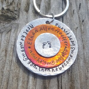 May include: A silver metal keychain with a round, two-toned disc. The disc has an orange and yellow center with the text "Ethan Allen Hoeffner*" and "now he's my angel*" in a circular pattern. The outer ring is silver and has the text "first he was my best friend, then he became my son."