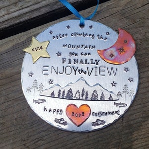 May include: A silver round ornament with a mountain scene, a crescent moon, and stars. The text reads "after climbing the mountain you can finally enjoy the view". There is a heart with the year 2022 and the words "happy retirement".