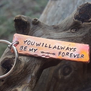 以下が含まれることがあります： 「You will always be my forever」という文字が刻まれた銅製のキーホルダー。キーホルダーはハンマーで叩いたような質感で、シルバーのキーリングが付いています。