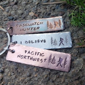 Op de afbeelding: Drie metalen sleutelhangers met verschillende ontwerpen. De bovenste sleutelhanger is koperkleurig en heeft de tekst "Sasquatch Hunter" met een silhouet van Bigfoot en dennenbomen. De middelste sleutelhanger is zilverkleurig en heeft de tekst "Ik geloof" met een silhouet van Bigfoot en dennenbomen. De onderste sleutelhanger is koperkleurig en heeft de tekst "Pacific Northwest" met een silhouet van Bigfoot en dennenbomen.