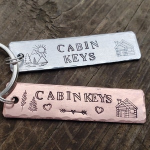 May include: Two silver and copper keychains with the text "CABIN KEYS" engraved on them. Both keychains feature a small cabin illustration and other decorative elements.