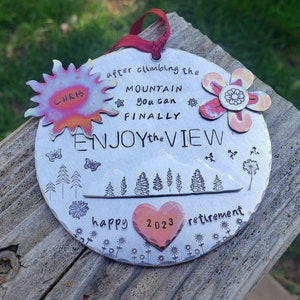 May include: A silver metal ornament with a mountain scene, trees, butterflies, and flowers. It says "after climbing the mountain you can finally enjoy the view" and "happy 2023 retirement".