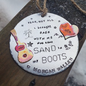 May include: A round metal ornament with a distressed finish. It features a guitar, palm tree, waves, cowboy boots, and a heart with a sun and a motorcycle. The text reads "Yeah, but all I brought back with me was some sand in my boots Morgan Wallen".