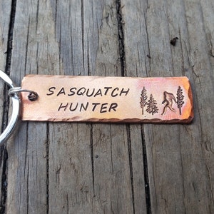 Op de afbeelding: Een koperkleurige sleutelhanger met de tekst "Sasquatch Hunter" en een silhouet van een sasquatch die door een bos loopt.