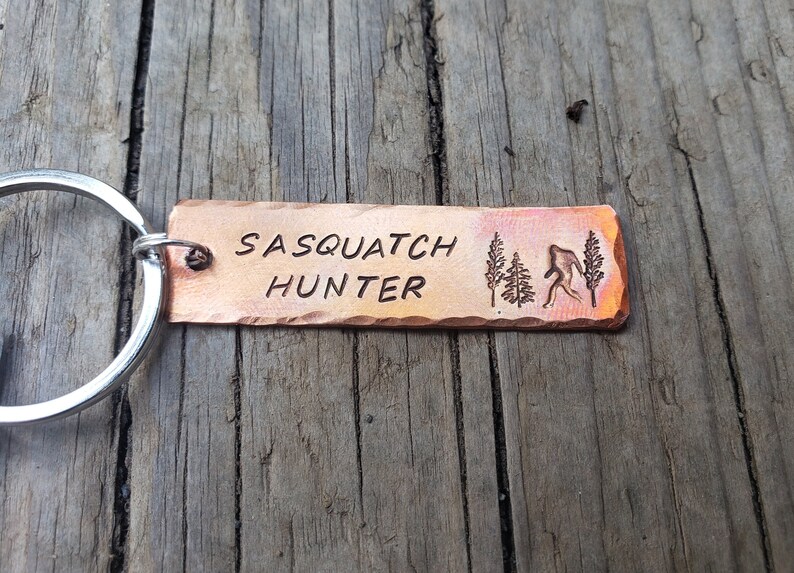 Op de afbeelding: Een koperkleurige sleutelhanger met de tekst "Sasquatch Hunter" en een silhouet van een sasquatch die door een bos loopt.