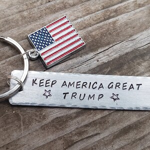 May include: A silver keychain with a small American flag charm and a rectangular tag that reads "KEEP AMERICA GREAT TRUMP" with a star on either side of the text.