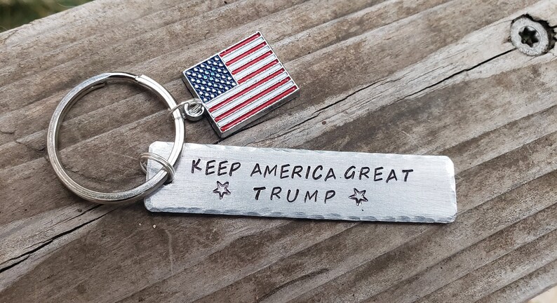 May include: A silver keychain with a small American flag charm and a rectangular tag that reads "KEEP AMERICA GREAT TRUMP" with a star on either side of the text.